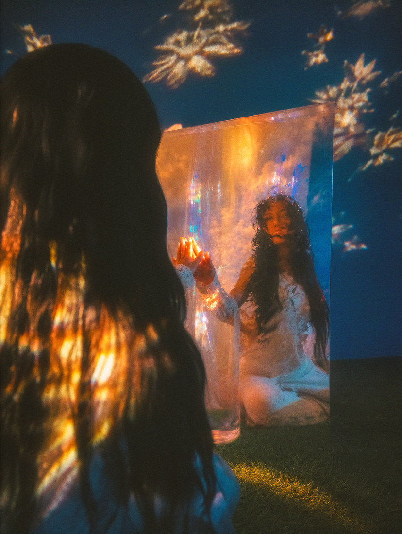 Ambyr seen from behind and reflected in a mirror under golden floral projections.