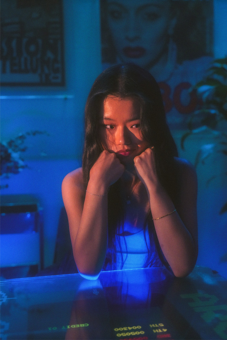 Ambyr leaning on a glass table with her hands near her face under blue light.