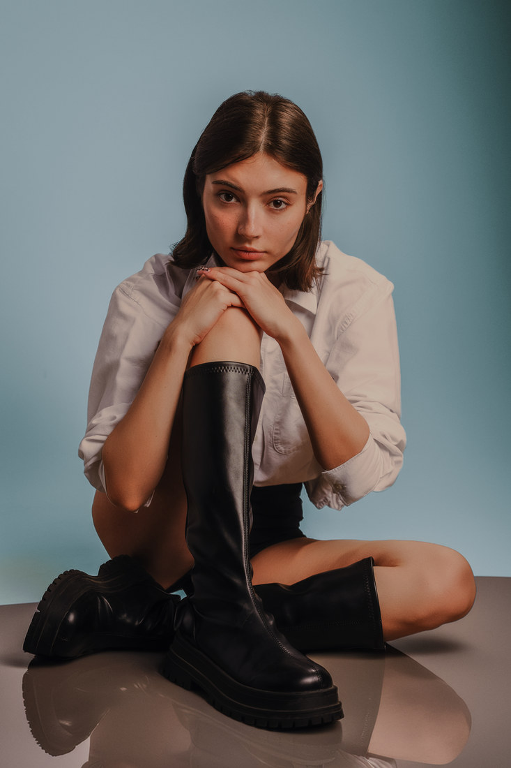Alla seated on the studio floor in a white shirt and black boots.