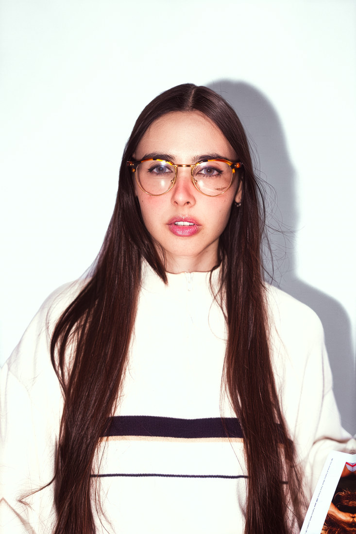 Studio portrait of Mima in glasses and a cream top with a dark horizontal stripe.