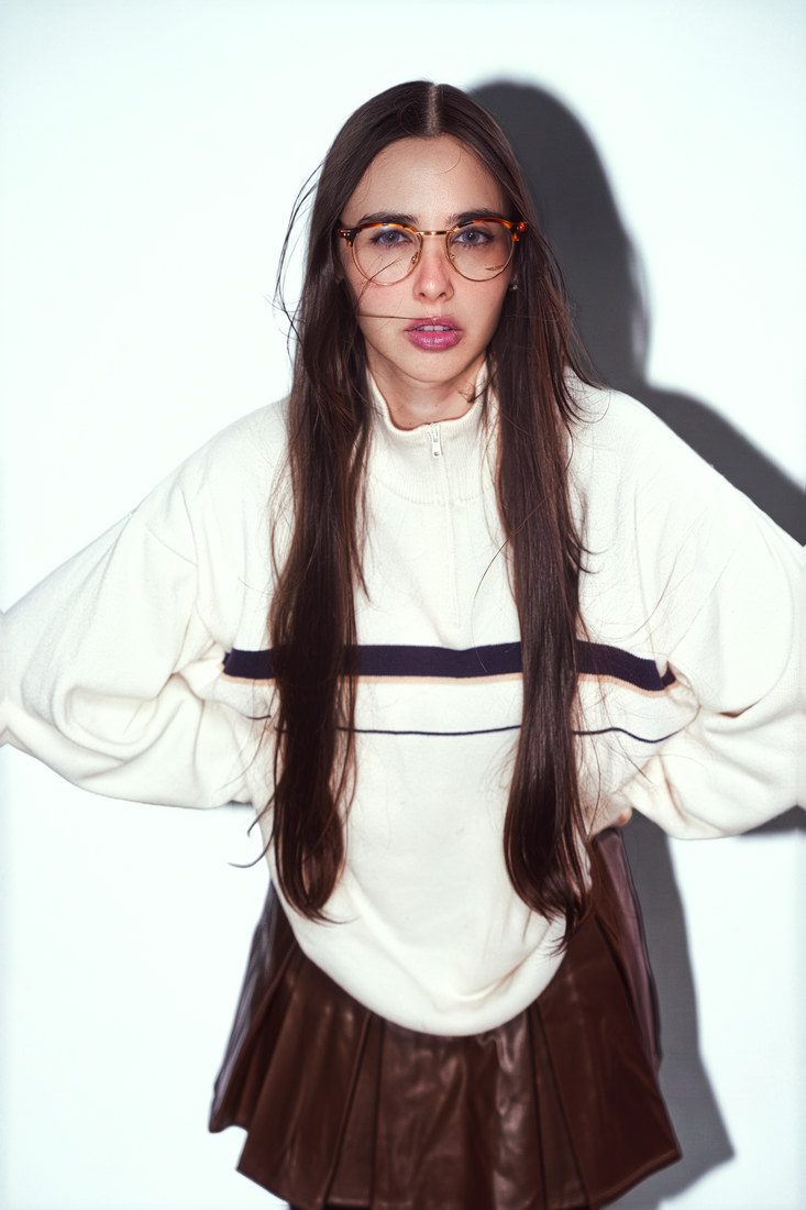 Mima in glasses, a cream top and a dark skirt, standing with her hands at her hips.