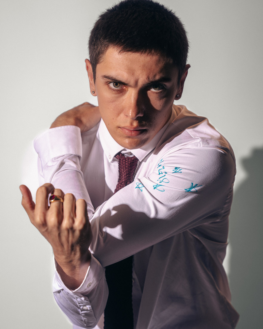 Model in a pale shirt and tie adjusting a cuff against a light background.