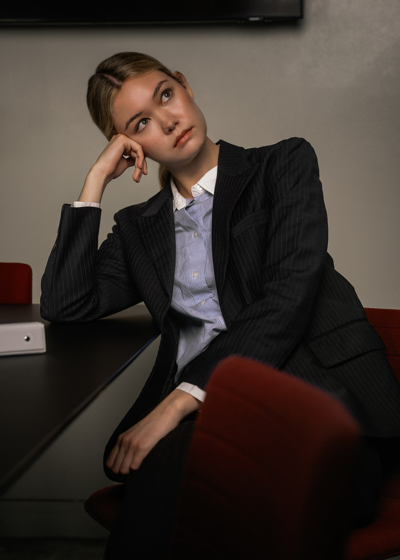 Model in a dark suit seated in a red office chair with her head resting on one hand.