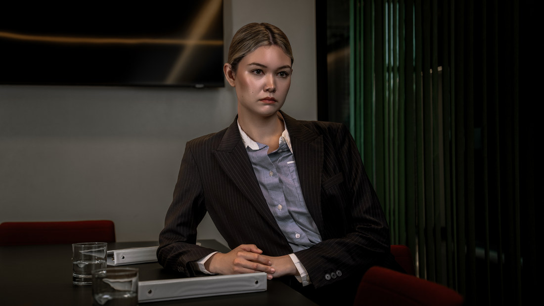 Model in a suit seated at a meeting-room table beside a drinking glass.